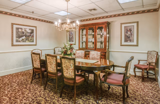 A formal dining room with a wooden oval dining table surrounded by eight upholstered chairs with floral patterns. A chandelier hangs above the table, and a wooden china cabinet with glass doors displays plates and glassware. The room has patterned carpet, wallpaper with a decorative border, and framed floral artwork on the walls.