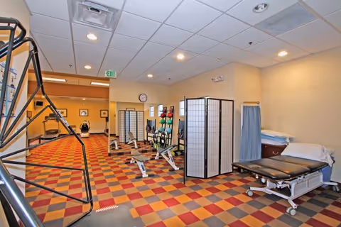 A spacious physical therapy or rehabilitation room with colorful checkered flooring, exercise equipment including rowing machines, therapy balls on racks, privacy screens, and an adjustable treatment table with pillows and a curtain for privacy.