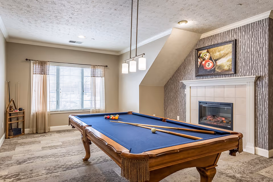 A well-lit game room featuring a pool table with blue felt, two pool cues, and a set of billiard balls arranged on the table. The room has a large window with sheer curtains, a modern ceiling light fixture with three lights, and a fireplace with a framed picture of billiard balls above it. The walls are painted beige with a textured accent wall behind the fireplace.