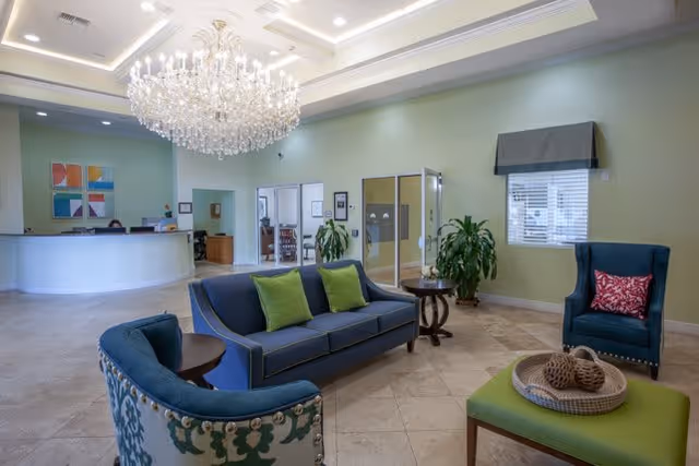 A spacious and well-lit reception area with a large crystal chandelier hanging from the ceiling. The room features a curved white reception desk with a person seated behind it, a blue sofa with green cushions, a blue armchair with a red patterned pillow, a green ottoman with decorative items on top, and a small round wooden table. The walls are painted light green, and there are large windows with blinds and plants placed around the room.