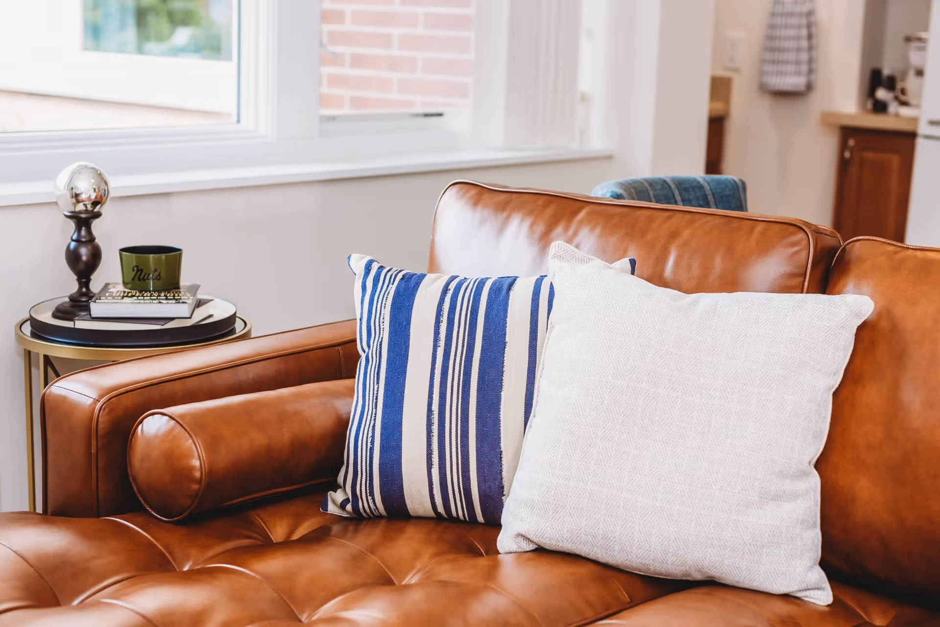 Close-up of a brown leather couch with two decorative pillows, one with blue and white stripes and the other in a light textured fabric. Next to the couch is a small round side table with a lamp, a green cup labeled 'Nuts', and some books. A window and part of a kitchen area are visible in the background.