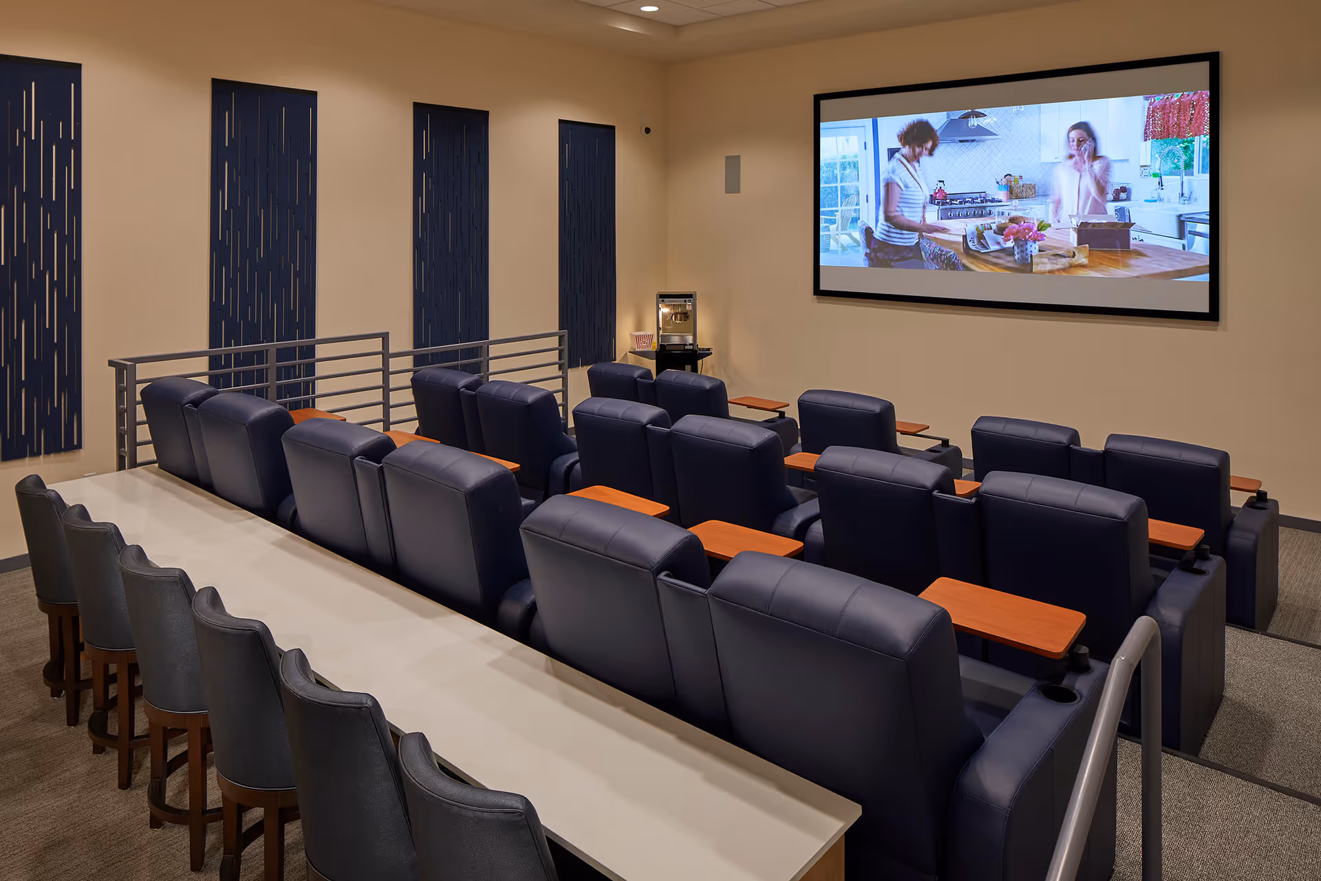 A small theater room with two rows of black leather recliner chairs equipped with wooden armrest trays, facing a large screen showing two women in a kitchen. Behind the recliners is a long counter with several high chairs. The walls are beige with decorative blue panels.