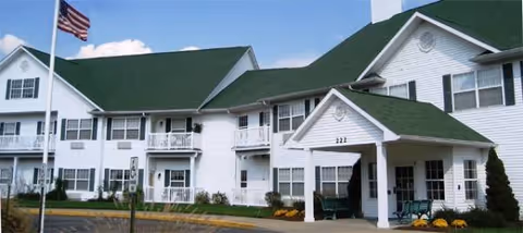 Exterior view of Sycamore Manor, a two-story white building with green roof shingles, multiple windows, balconies, and an American flag flying on a flagpole in front. The entrance has a covered porch with seating and landscaping with yellow flowers.