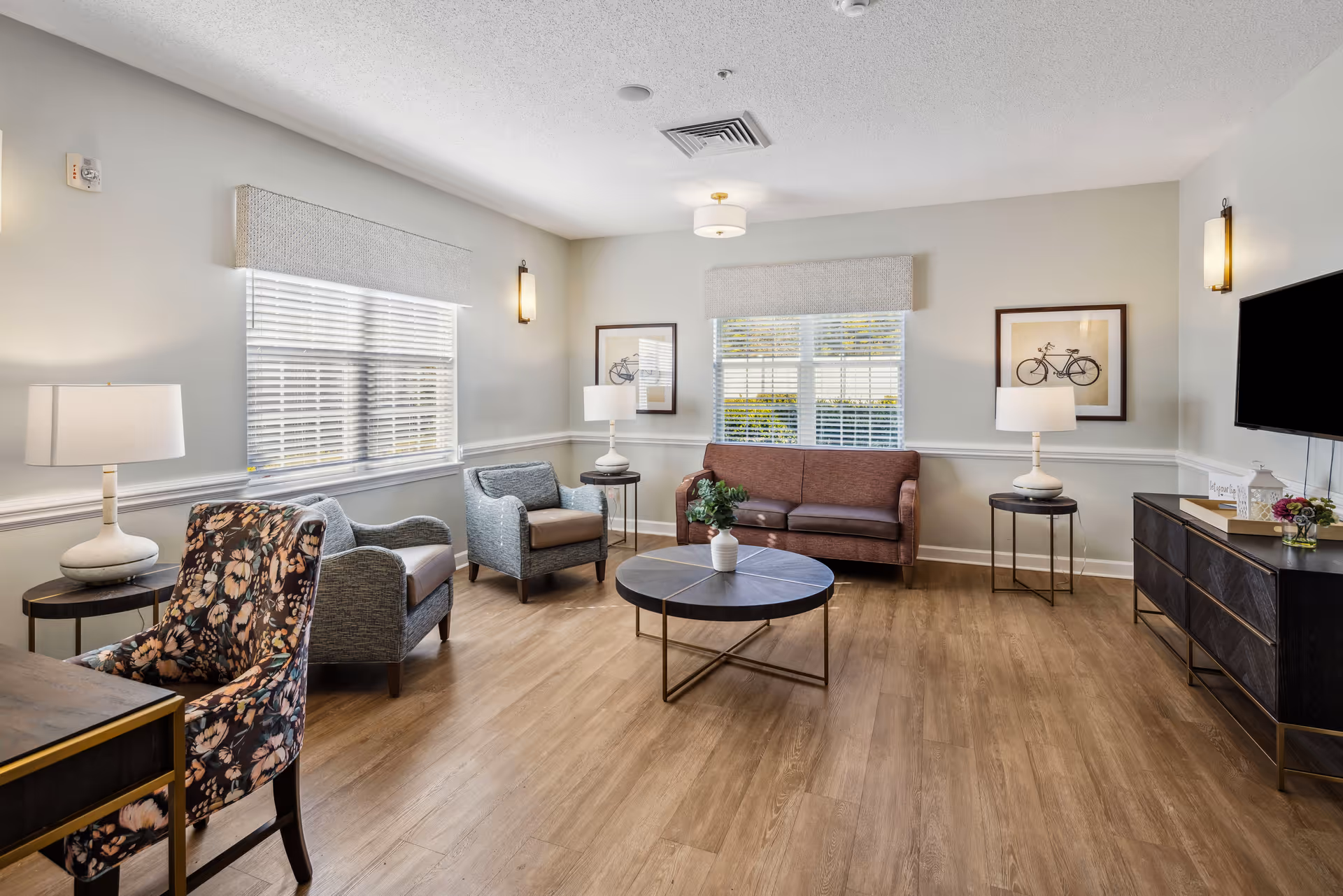 Bright communal living room with sofas and armchairs arranged around a round coffee table, wood floors, windows with blinds, lamps, and wall art.