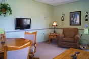 A cozy living room area with a wall-mounted flat screen TV, a wooden dining table with chairs, a brown upholstered armchair, a wooden side table with a lamp, and a framed picture on the light green wall. There is also a hanging plant in the corner.