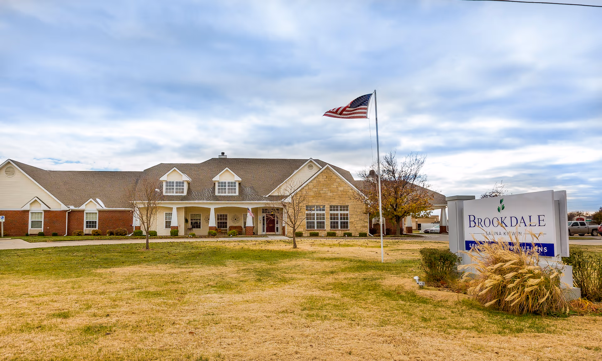 Front exterior of the Brookdale Salina Kirwin senior living building with an American flag on a flagpole and a sign on a grassy lawn.