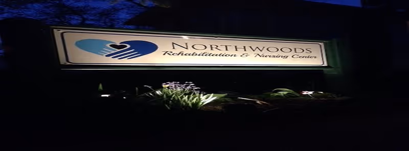 Illuminated sign at night for Northwoods Rehabilitation & Nursing Center with a logo of two hands forming a heart shape.