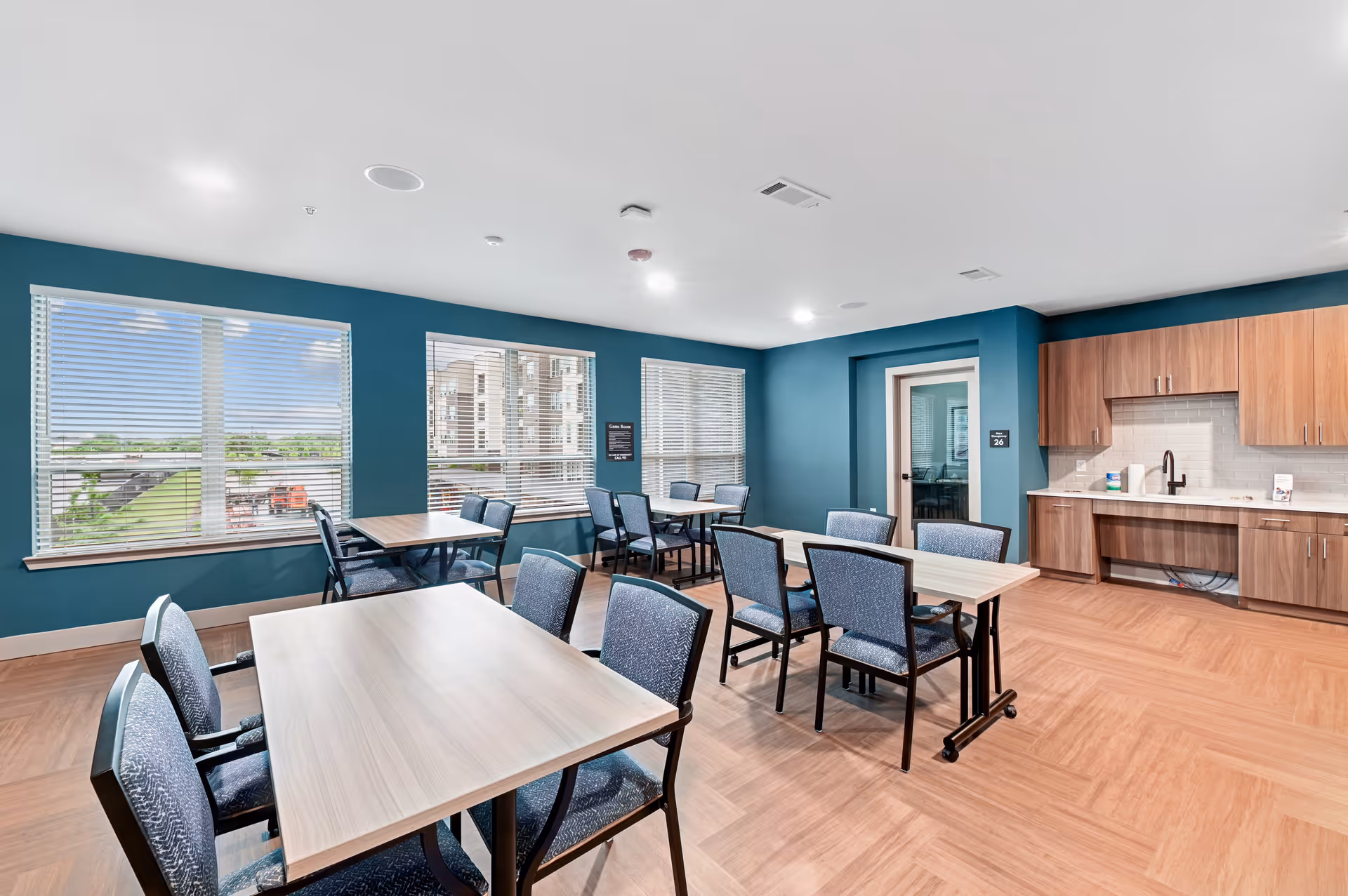 A bright and clean community room with several tables and chairs arranged for group seating. The room has teal walls, large windows with white blinds letting in natural light, and a kitchenette area with wooden cabinets and a sink. The floor is a light wood pattern, and there is a door leading to another room.