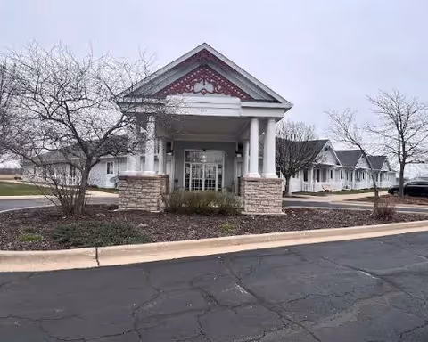 Front entrance of a single-story white senior living building with a covered porte-cochere supported by columns, leafless trees, and a paved driveway in the foreground.