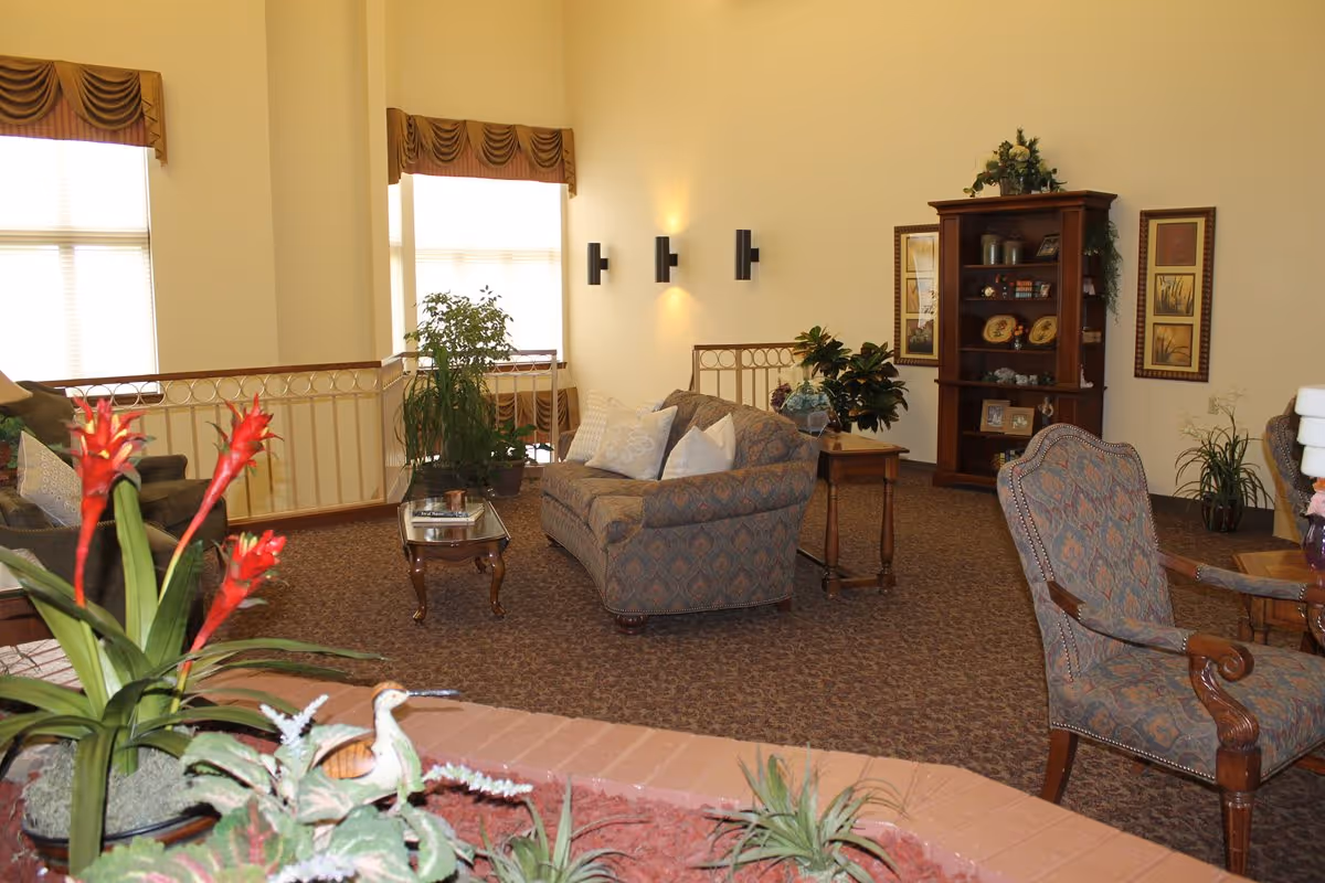 Sunlit common living area with patterned sofas and armchairs, plants, side tables, and a bookshelf.
