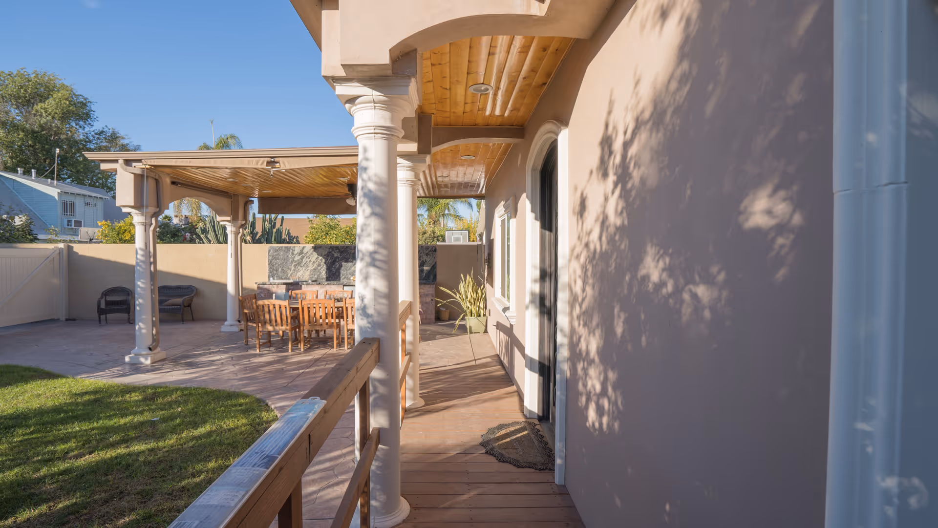 Outdoor patio area with a covered seating space featuring wooden chairs and a table, white columns supporting the roof, and a small grassy lawn adjacent to the patio. The scene is sunlit with shadows of trees cast on the wall of the building.
