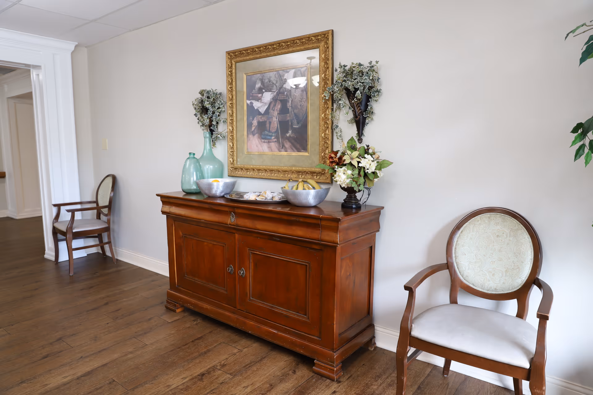 A hallway area with a wooden sideboard against a light-colored wall. On top of the sideboard are decorative items including two large green glass bottles, two metal bowls with fruit, and a floral arrangement. Above the sideboard hangs a framed painting with a gold ornate frame. To the right of the sideboard is a wooden chair with a cushioned seat and backrest, and another similar chair is visible further down the hallway. The floor is wooden and the space is well-lit.