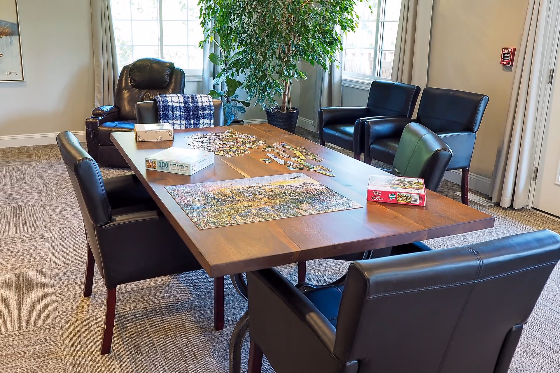 Sunlit common room with a wooden activity table surrounded by leather chairs and a partially completed jigsaw puzzle.