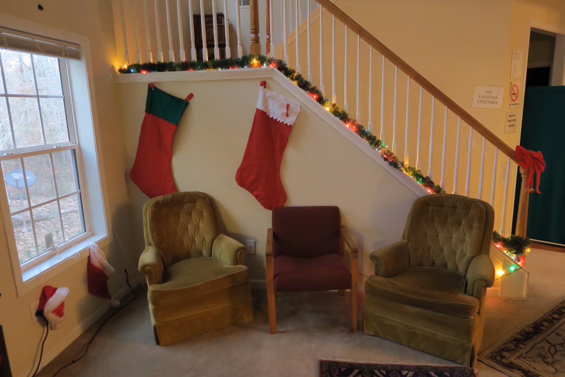 A cozy corner of a living room with two green upholstered armchairs and a smaller maroon chair between them. The wall behind the chairs is decorated with two large Christmas stockings hanging from a staircase railing adorned with green garland and colorful Christmas lights. A window to the left shows a view of trees outside, and a small Santa hat is placed on the windowsill. There are signs on the wall near the staircase, including one that says 'NO VISITORS UPSTAIRS'.