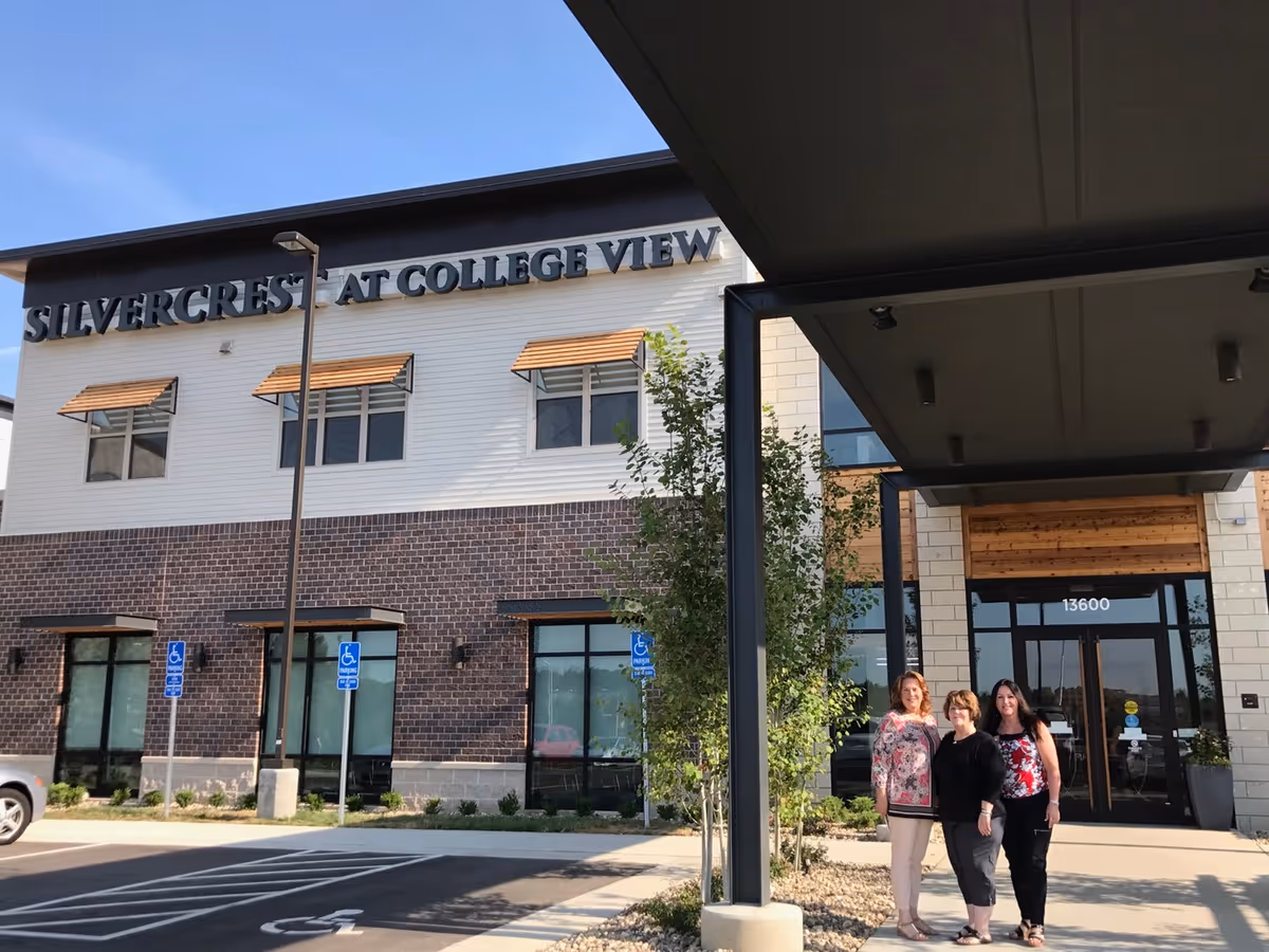 Exterior front view of Silvercrest at College View Senior Living building with three women standing near the entrance under a covered walkway. The building has a brick and white facade with large windows and the address number 13600 above the door.