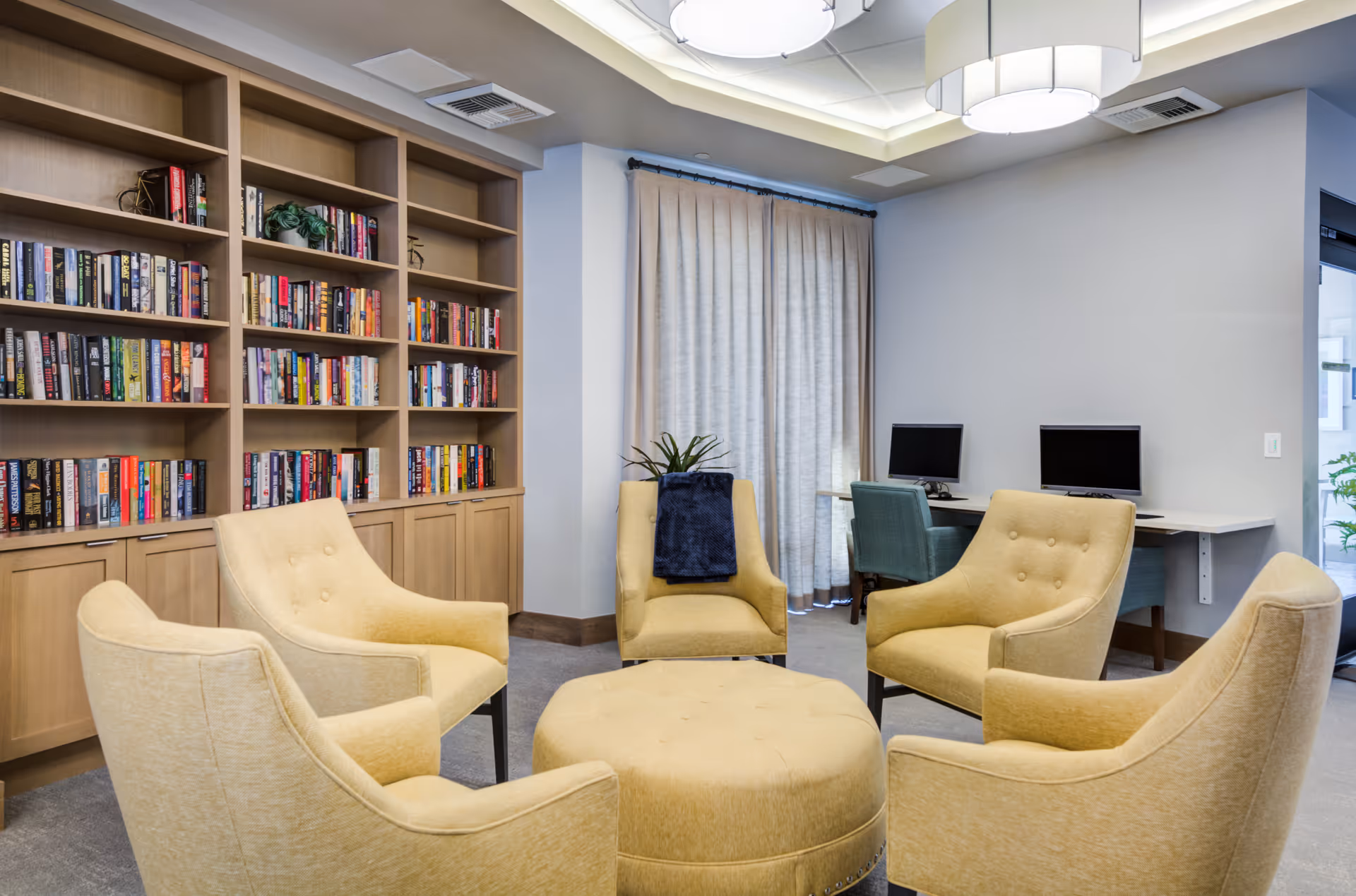 Cozy communal lounge with a round arrangement of yellow armchairs and an ottoman facing built-in bookshelves, with a small computer station in the background.