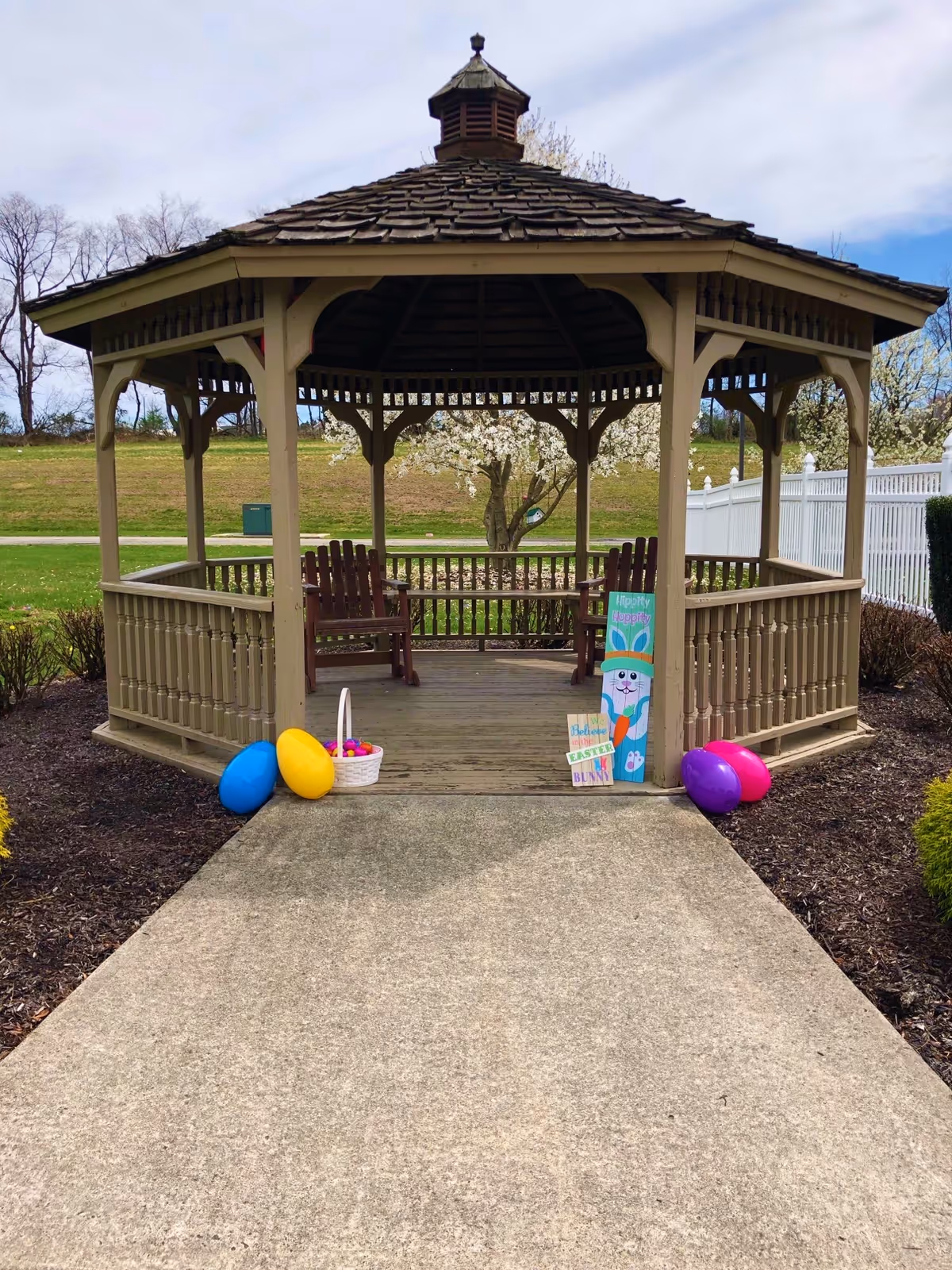 Wooden gazebo in a grassy outdoor area decorated with oversized colorful Easter eggs and a bunny sign.
