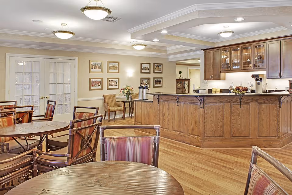 A cozy dining area with round wooden tables and chairs featuring striped cushions. The room has a wooden floor and a wooden counter with cabinets above it. The walls are decorated with framed pictures, and there are double glass doors on one side. Ceiling lights provide warm illumination.