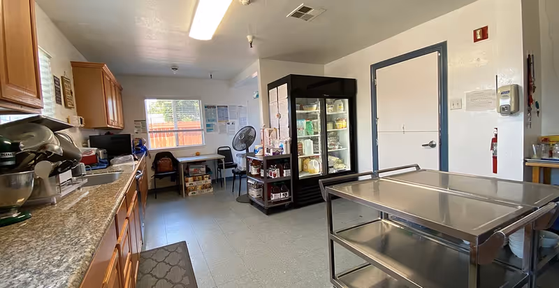 A commercial-style kitchen/food prep room with countertops and cabinets on the left, a glass-front refrigerator, stainless steel serving cart and a small table under a window.