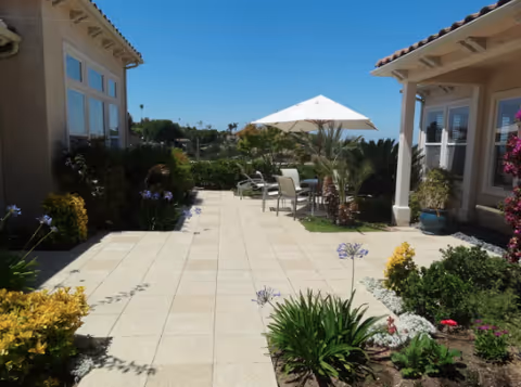 Outdoor patio area between two buildings with tiled flooring, surrounded by various green plants and flowers. There is a table with chairs under a large white umbrella in the center, and the sky is clear and blue.