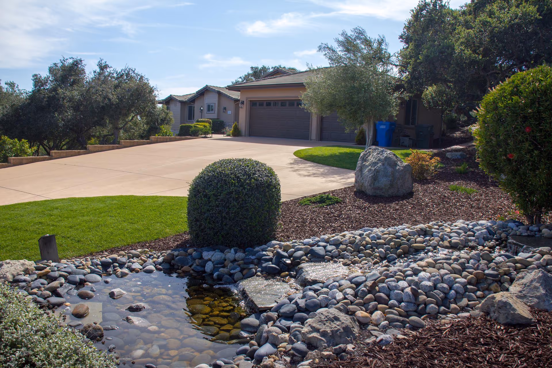 Landscaped front yard and driveway with a rock-lined water feature, trimmed shrubs, and a garage in the background.