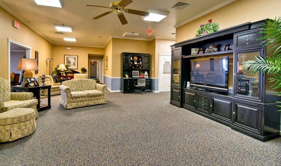 A spacious living room area in a senior living facility with patterned carpet flooring, beige walls, and ceiling fans. The room features multiple upholstered chairs and a loveseat, a large black entertainment center with a flat-screen TV, a desk with a computer, several table lamps, framed artwork on the walls, and decorative plants.