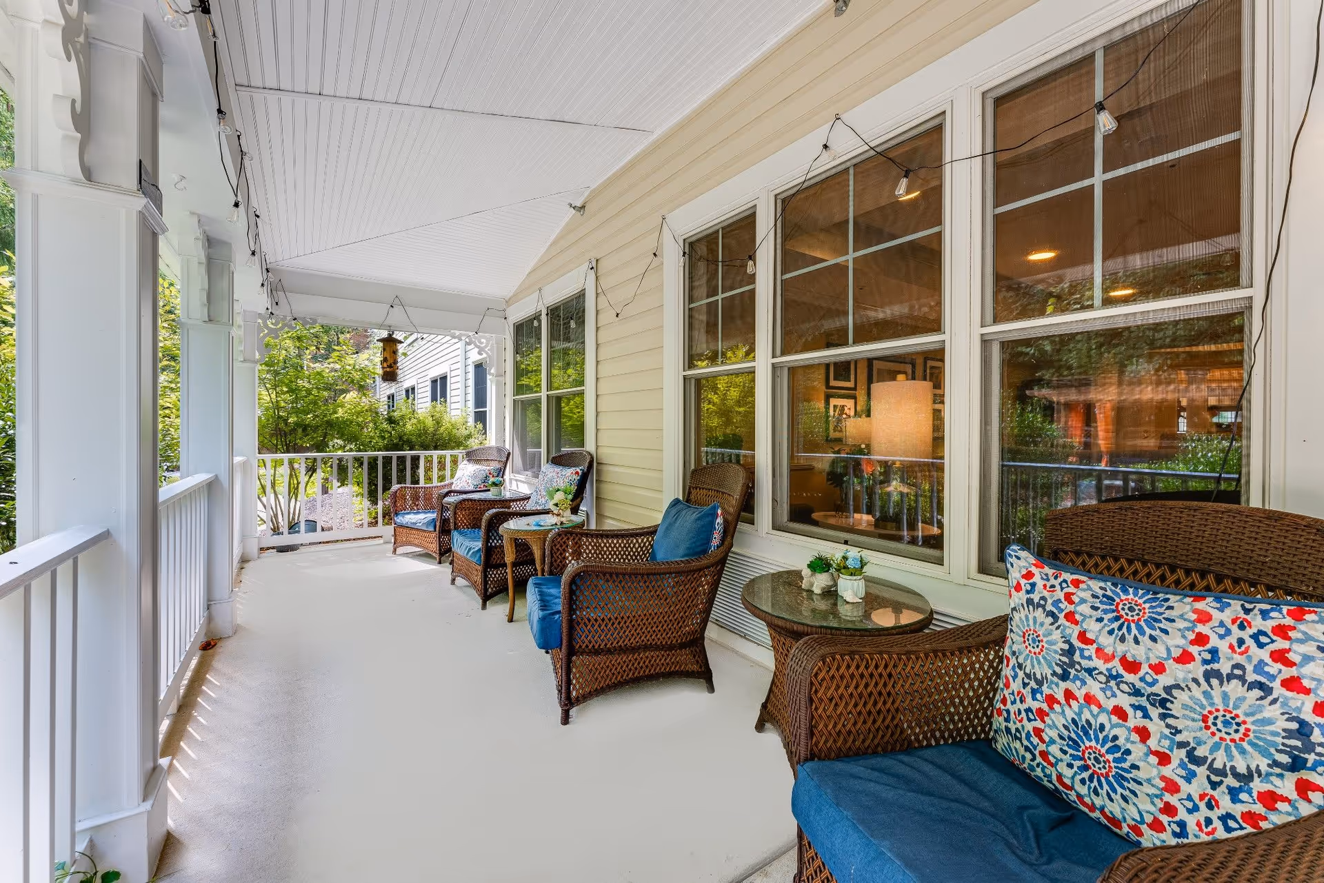 Covered outdoor porch area with wicker chairs and small tables. The chairs have blue cushions and colorful patterned pillows. String lights hang from the ceiling, and there are large windows on the right side reflecting indoor lighting and decor. Greenery and neighboring buildings are visible beyond the porch railing.