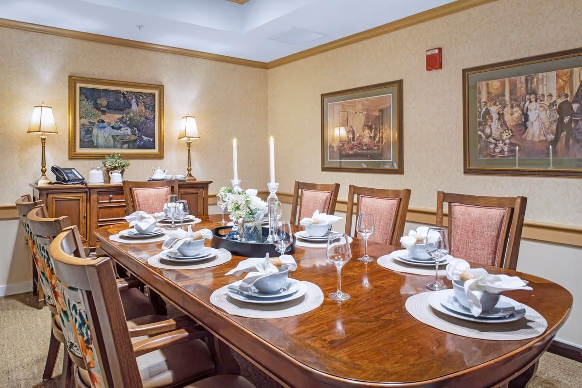 A formal dining room with a large wooden dining table set for eight people. The table is arranged with white plates, bowls, napkins, wine glasses, and a centerpiece with flowers and two lit candles. The room has beige wallpaper with framed paintings on the walls and a wooden sideboard with two lamps and a telephone.