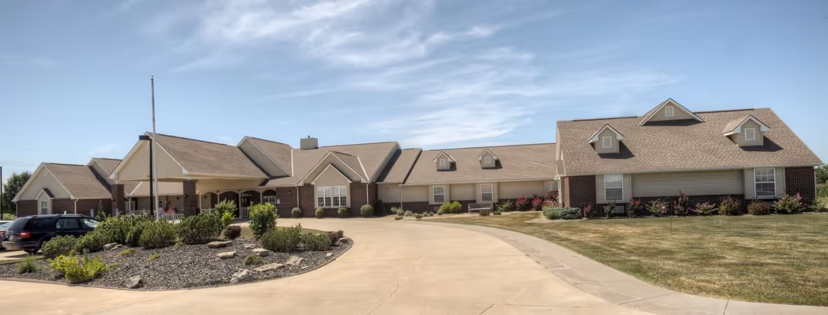 Exterior view of a single-story senior living facility building with a beige roof and brick accents, surrounded by a driveway and landscaped greenery under a partly cloudy sky.
