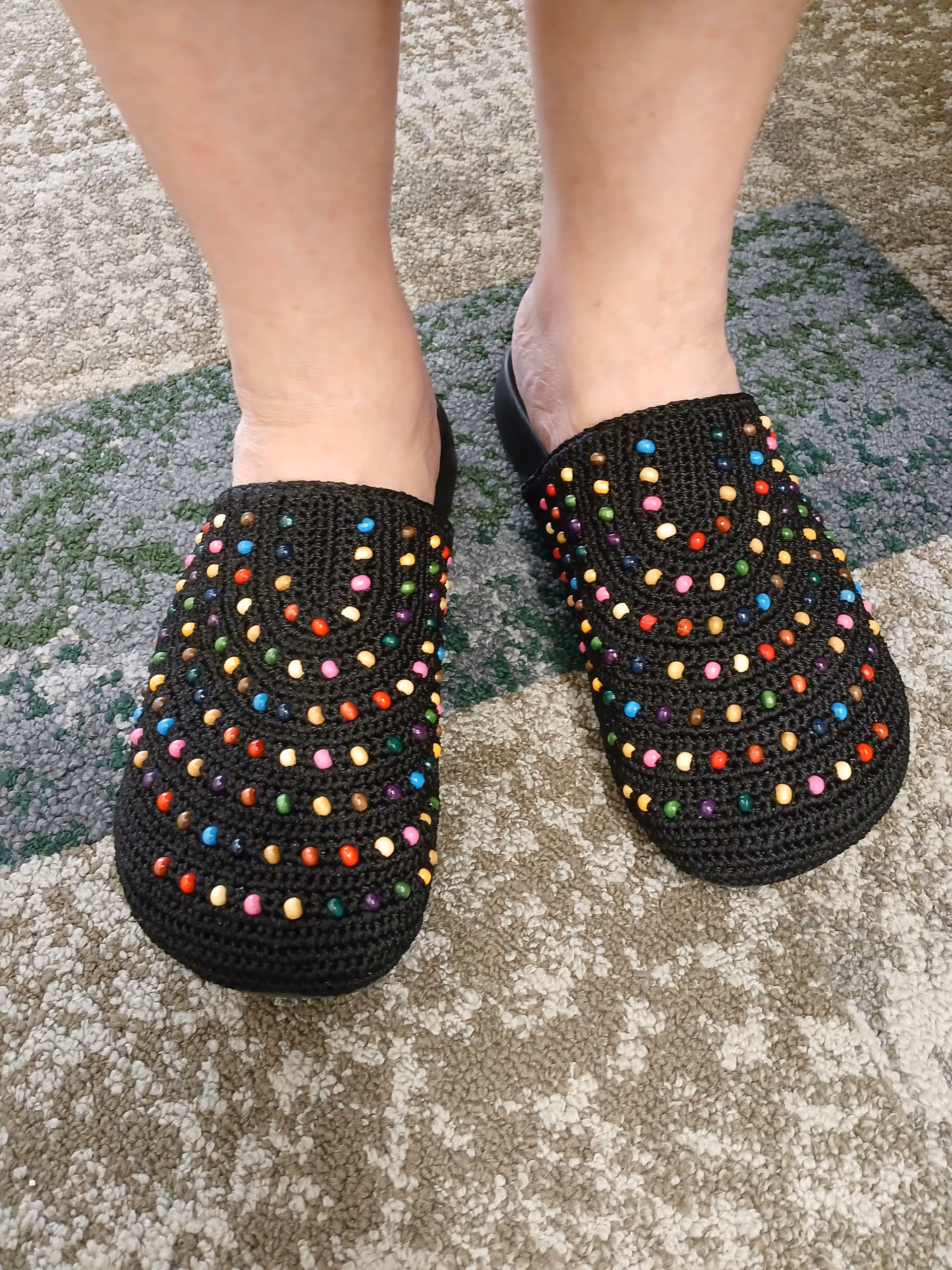 A pair of feet wearing black crocheted slippers decorated with colorful beads standing on patterned carpet.