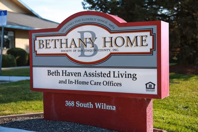 Outdoor sign for Bethany Home Society of San Joaquin County, Inc. with text indicating Beth Haven Assisted Living and In-Home Care Offices, located at 368 South Wilma, set on a red base with grass and building in the background.
