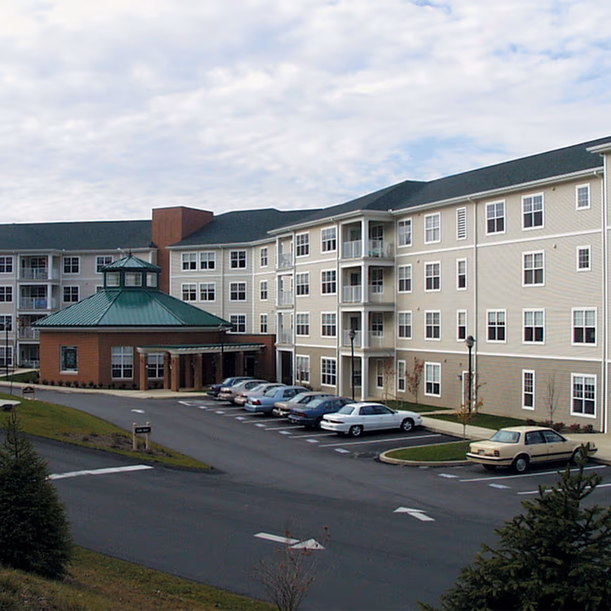 Exterior view of a multi-story senior living facility with beige siding and white trim. The building features multiple windows, balconies, and a green-roofed entrance area. Several cars are parked in the parking lot in front of the building under a cloudy sky.