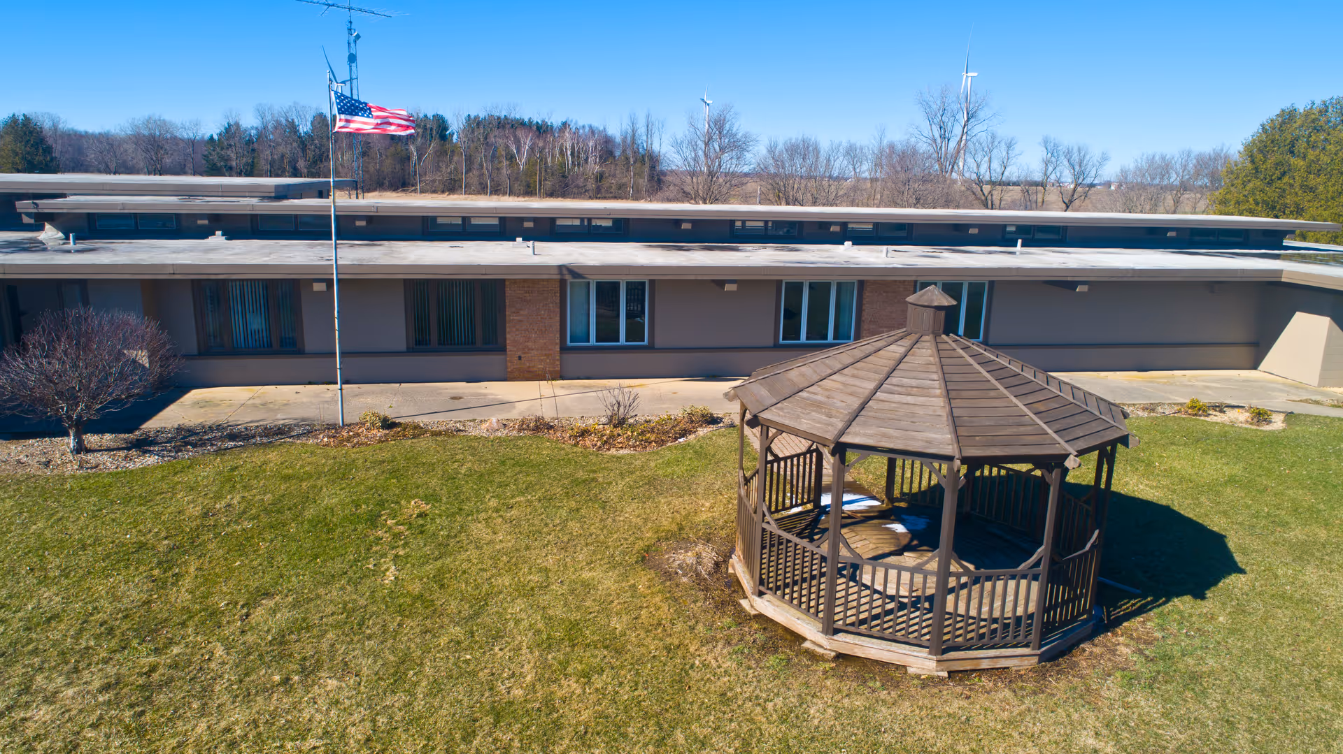 A grassy courtyard with a wooden gazebo and an American flag in front of a single-story building.
