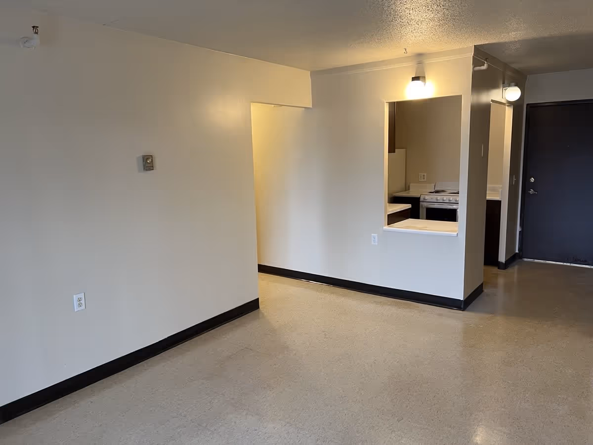 Empty interior room with beige walls and floor, a small kitchen area with a stove and countertop visible through a pass-through window, and a dark-colored door in the background.