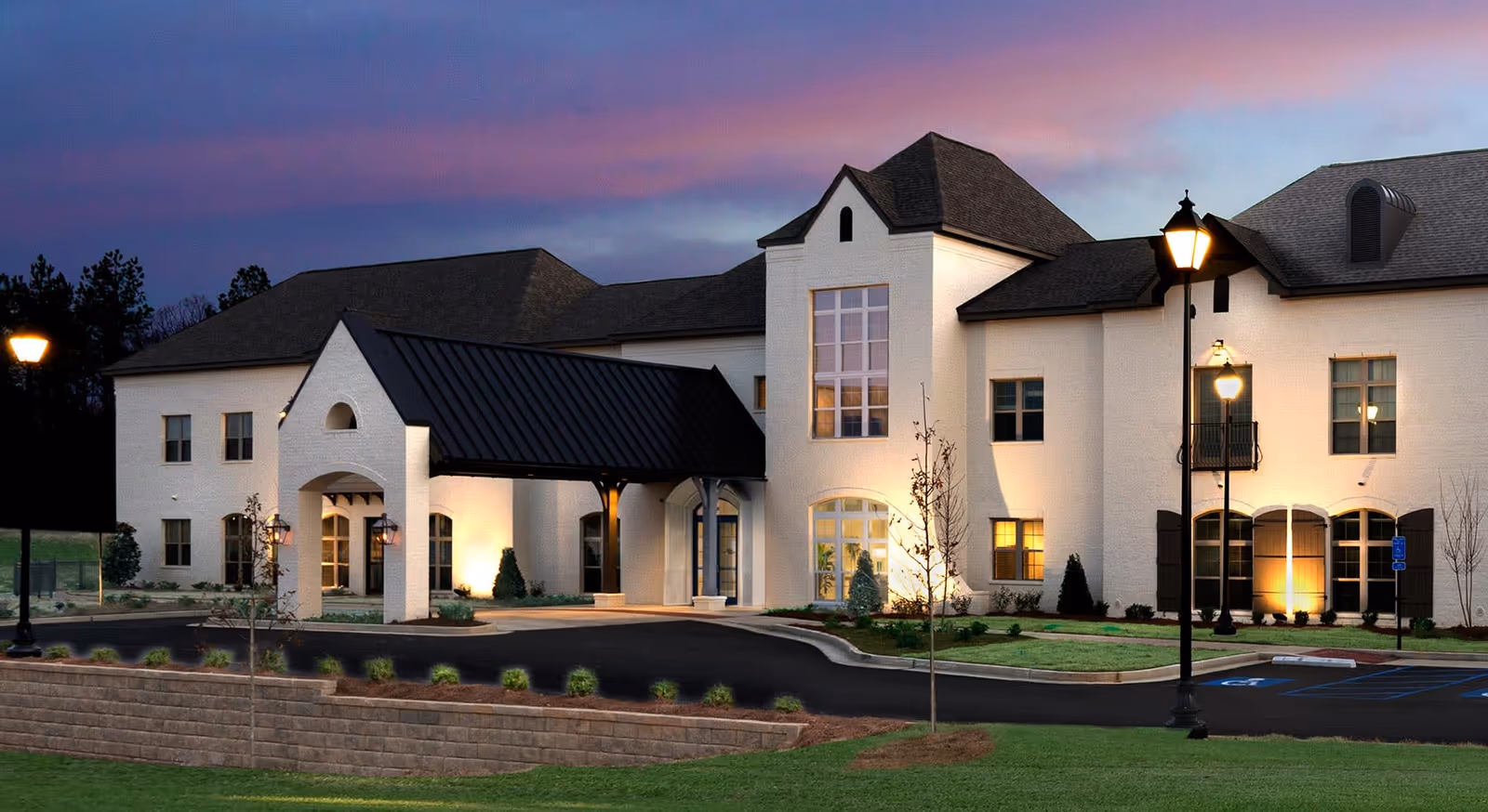 Exterior view of The Blake at Woodcreek Farms facility at dusk, showing a large white building with multiple windows, a covered entrance with a black roof, illuminated by outdoor lamps and surrounded by landscaped greenery and a parking area.