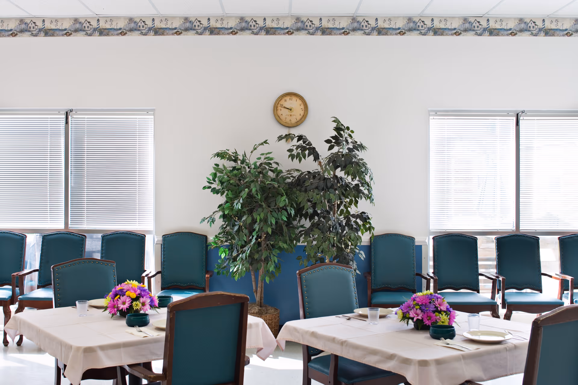A bright dining room with two tables covered in white tablecloths, each set with plates, utensils, and a small bouquet of purple and yellow flowers. There are multiple teal upholstered chairs around the tables and along the wall. Two large windows with blinds let in natural light. A clock hangs on the white wall above two large green potted plants. A decorative border runs along the top of the wall.