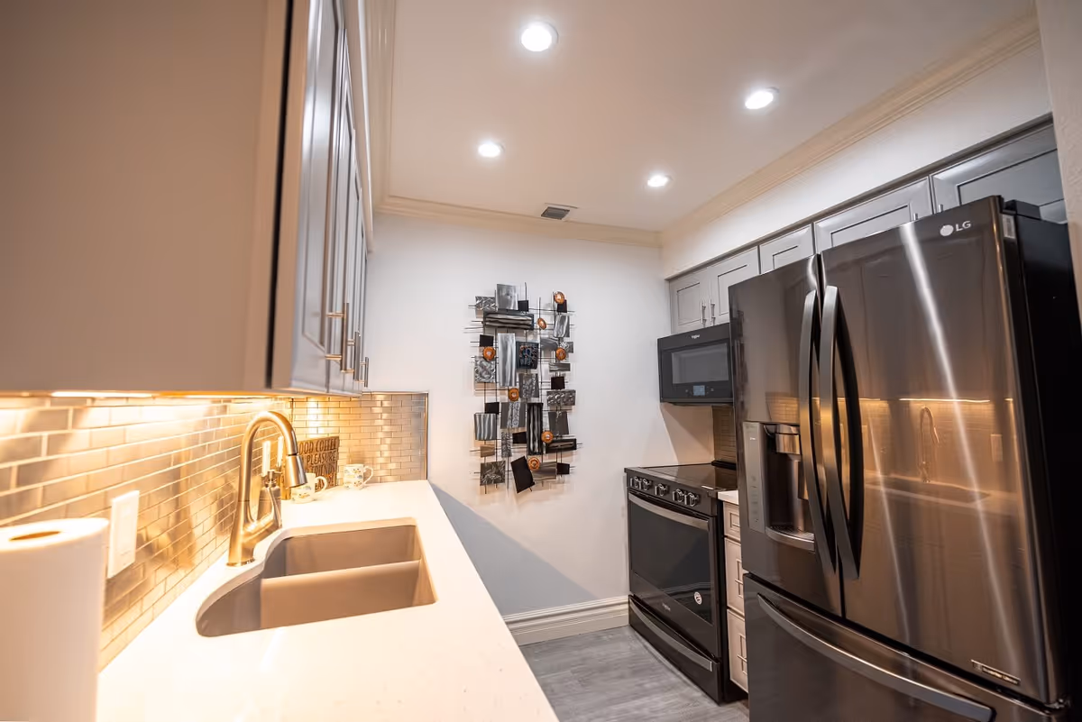 Modern kitchen with stainless steel refrigerator, black stove, microwave, white countertops, and under-cabinet lighting. There is a double sink with a gold faucet and decorative wall art on the far wall.
