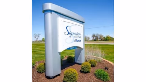 Outdoor view of a large sign for Signature HealthCARE of Muncie, placed in a landscaped area with bushes and grass under a clear blue sky.