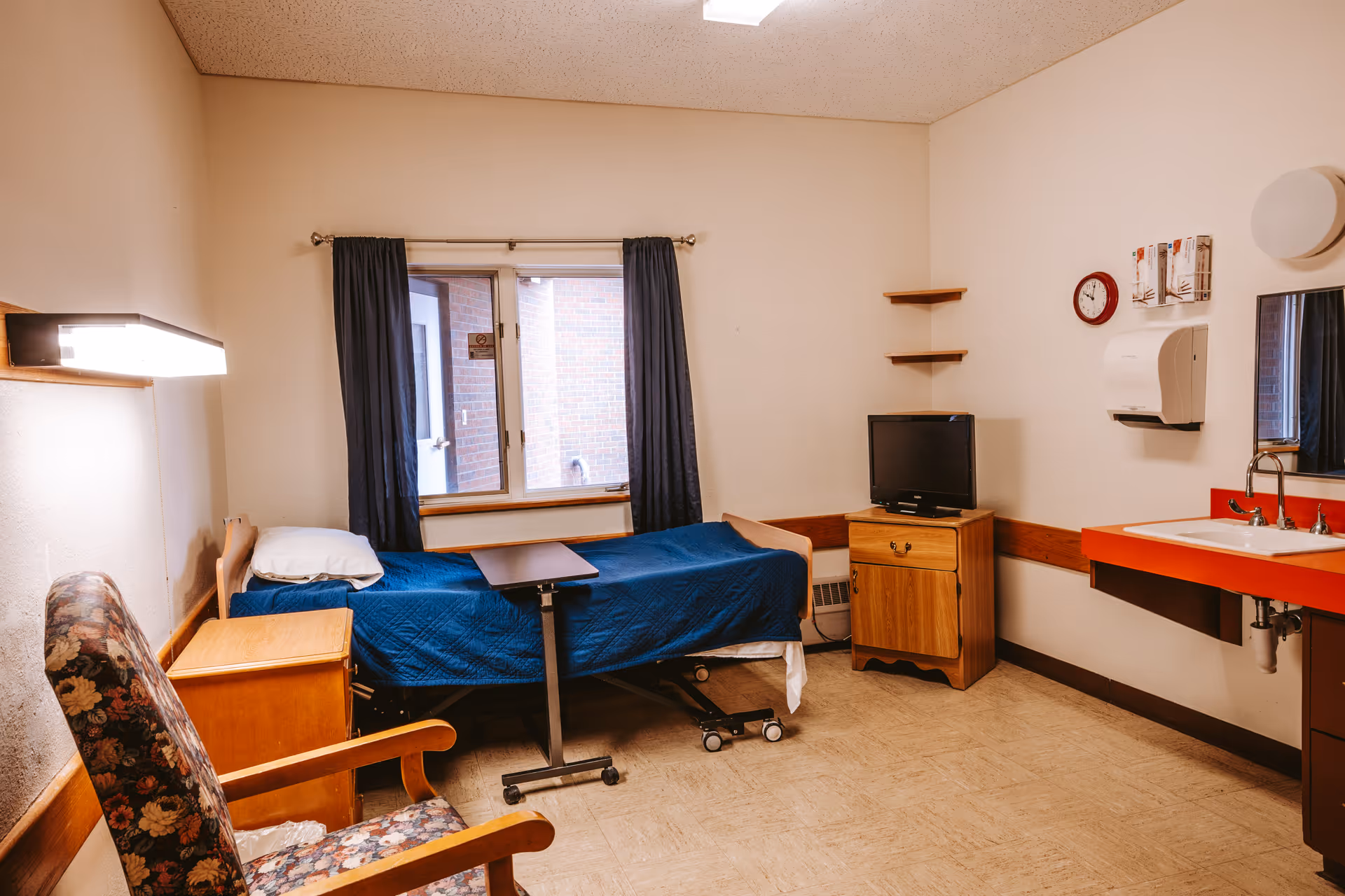 A small, simple patient room in a healthcare facility with a single bed covered in a blue blanket, a wooden nightstand, a floral upholstered chair, a small TV on a wooden cabinet, a wall-mounted sink with a red countertop, a mirror, and a window with dark curtains.