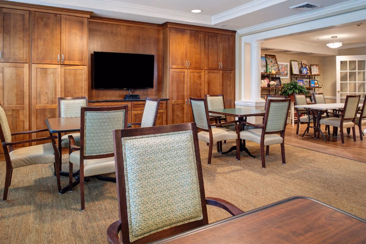 A well-lit common area with multiple wooden tables and cushioned chairs arranged on a carpeted floor. The back wall features wooden cabinets and a mounted flat-screen TV. In the background, there is an open space with more tables and chairs, decorative plants, and artwork displayed on shelves.