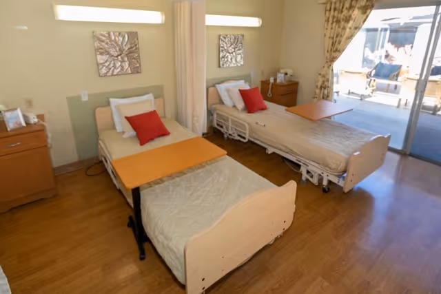 A bright room with two hospital-style beds, each with a beige quilted blanket and a red pillow. Each bed has a wooden overbed table. There are two wooden nightstands, one on each side of the beds, with a telephone and other small items. The room has wooden flooring and a large sliding glass door leading to an outdoor patio area with chairs. The walls are light-colored with two decorative wall hangings above the beds.