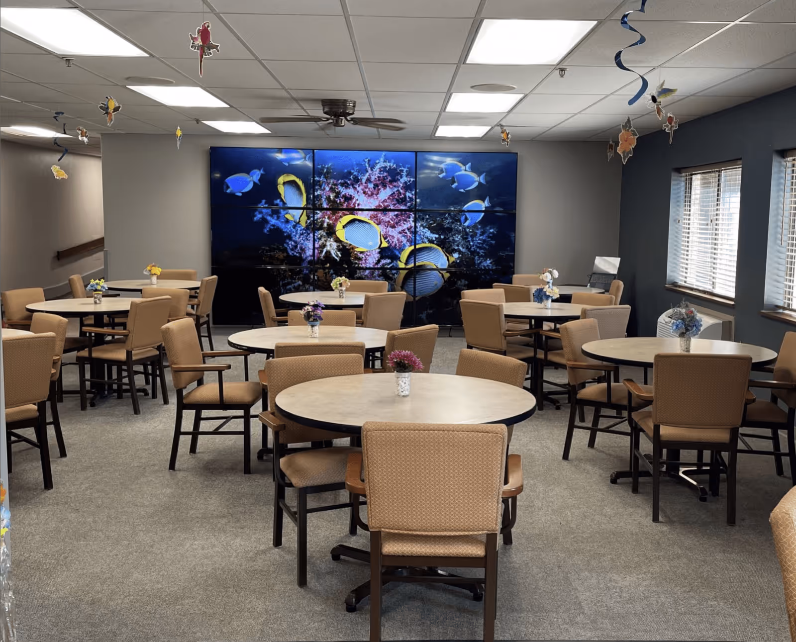 A senior living facility dining room with multiple round tables and beige cushioned chairs. Each table has a small flower arrangement. The room has a large wall-mounted screen displaying an underwater scene with colorful fish. The ceiling has fluorescent lights and a ceiling fan, and decorative hanging ornaments are suspended from the ceiling. Windows with blinds are on the right side of the room.