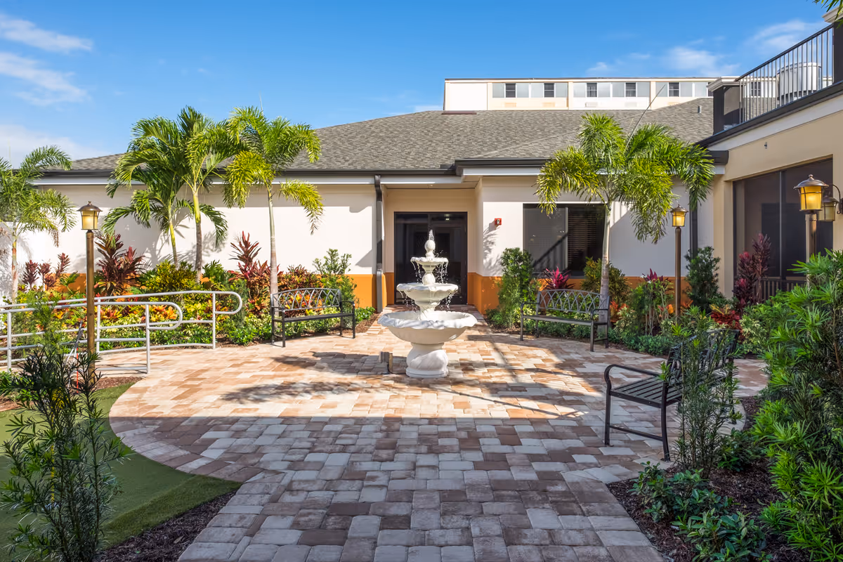 Outdoor courtyard area at Volante Senior Living of Melbourne featuring a central white tiered fountain surrounded by benches, palm trees, and various plants under a clear blue sky.