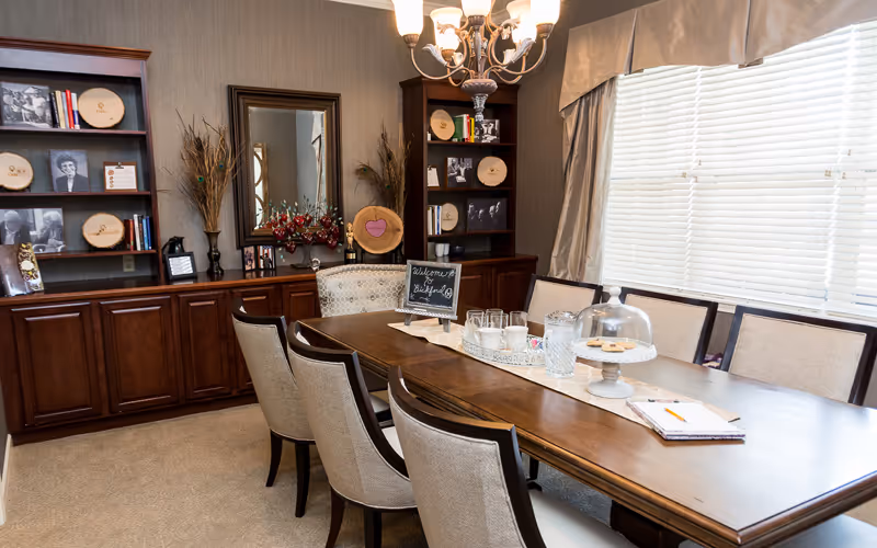 A well-lit dining room with a long wooden table surrounded by upholstered chairs. On the table, there is a tray with glasses, a pitcher, a covered plate with cookies, and a small chalkboard sign that says 'Welcome to Bickford'. The room has large windows with blinds and curtains, wooden cabinets with shelves displaying framed photos, books, and decorative items. A chandelier hangs above the table.
