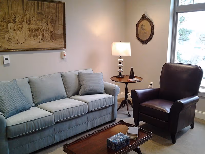 A cozy living room with a light blue sofa adorned with two cushions, a dark brown leather armchair, a wooden coffee table with a tissue box and books, and a round side table with a lamp and a metronome. The walls are decorated with two framed artworks, and there is a large window letting in natural light.