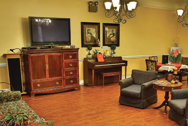 A cozy living room area with wooden flooring, featuring a large flat-screen TV on a wooden cabinet, a piano with a bench, two framed peacock paintings on the wall, two upholstered armchairs, a round wooden side table with magazines, and a dining table with chairs in the background. The walls are painted yellow with white trim, and there are two ornate chandeliers hanging from the ceiling.
