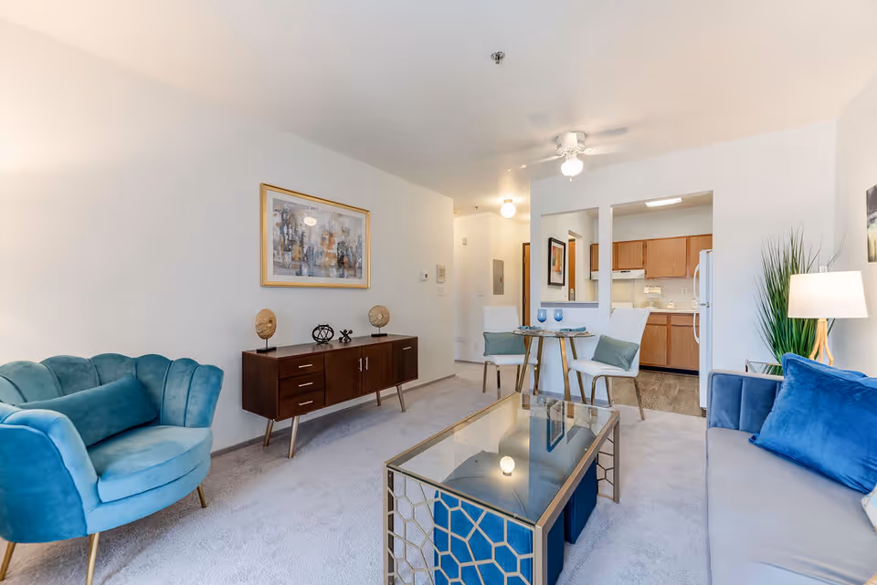 A bright and modern living room area with a blue velvet armchair and matching blue pillows on a gray sofa. There is a glass coffee table with a geometric gold frame in the center. Against the wall is a wooden sideboard with decorative items and a framed abstract painting above it. In the background, a small dining area with a round glass table and two white chairs is visible, along with a kitchen featuring wooden cabinets and white appliances. The room has light-colored walls and carpeted flooring.