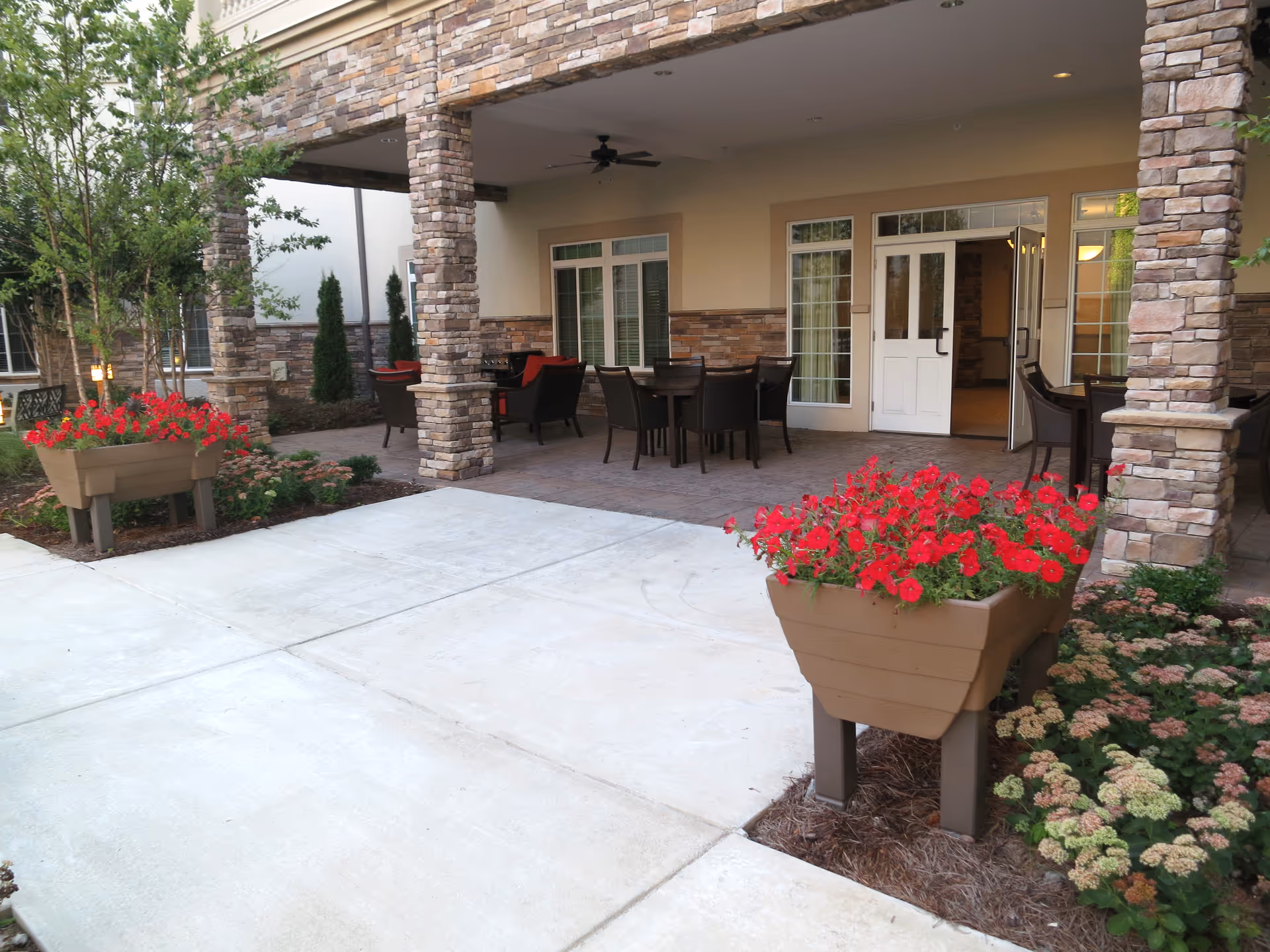 Outdoor covered patio area with stone pillars and beige walls, featuring several dark brown tables and chairs. Two large planters with vibrant red flowers flank the concrete walkway leading to the patio. Green shrubs and trees surround the area, creating a welcoming garden-like atmosphere.