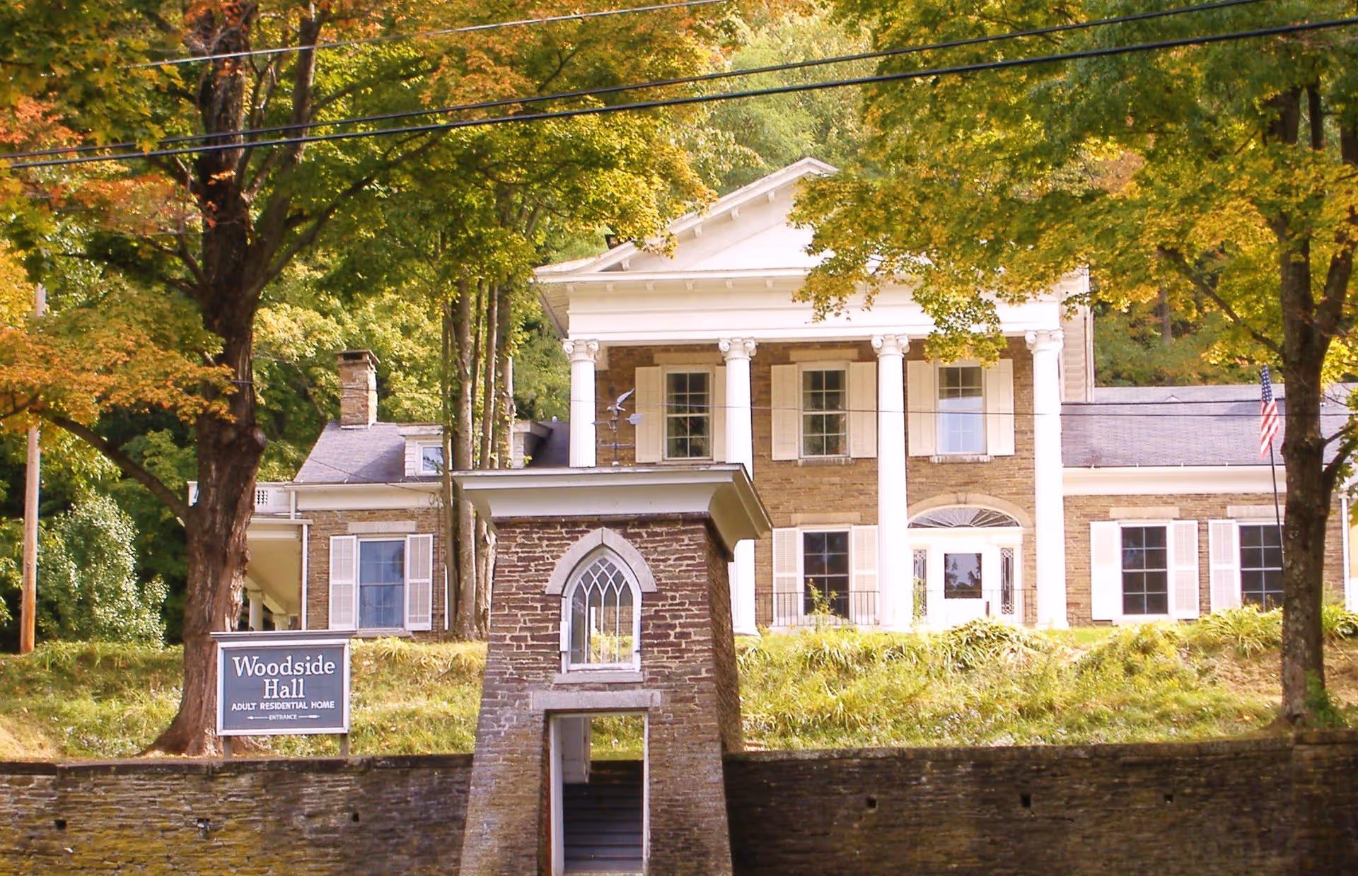 Front exterior view of Woodside Hall, an adult residential home, showing a large historic building with white columns, multiple windows with shutters, surrounded by trees with green and autumn-colored leaves.