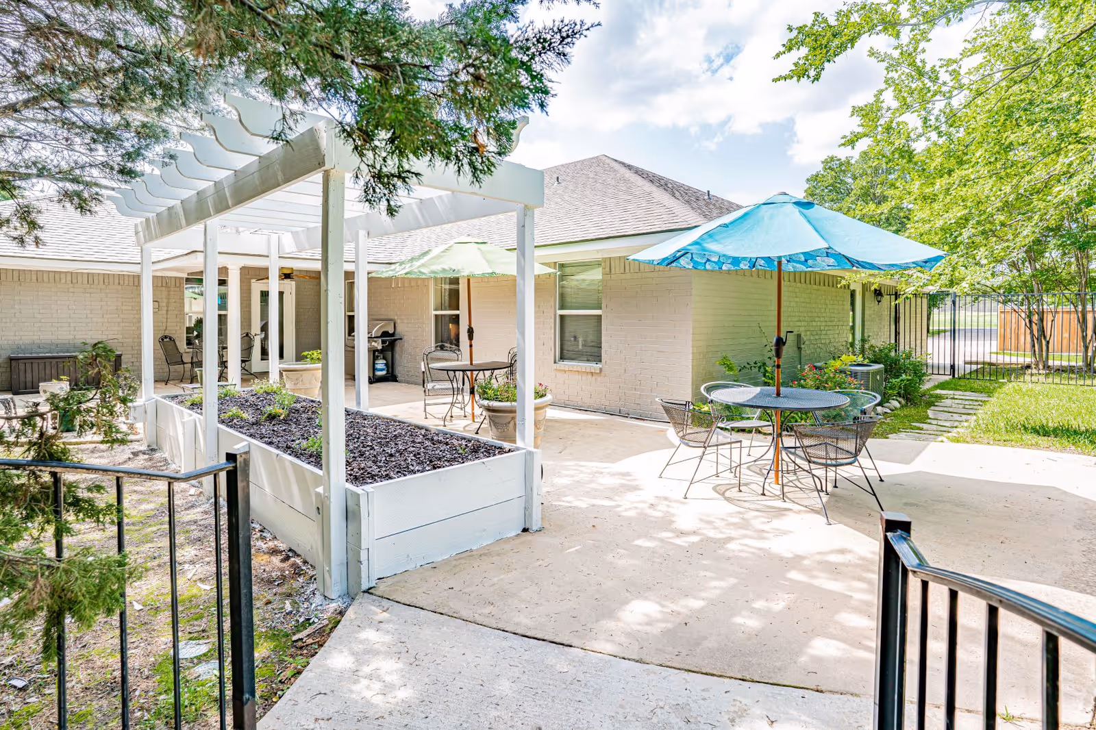 Sunny outdoor patio courtyard with tables, umbrellas, a pergola planter box and seating alongside a light-brick building.